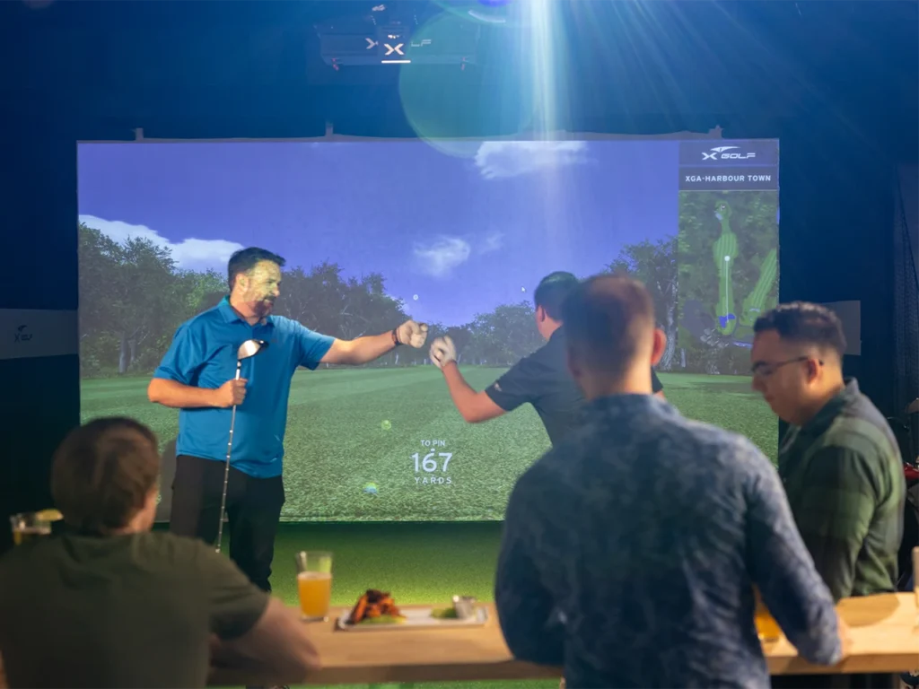 Two men sharing a fist bump in front of an indoor golf simulator during a bachelor party at X-Golf, with other attendees enjoying drinks and food at a table in the foreground.