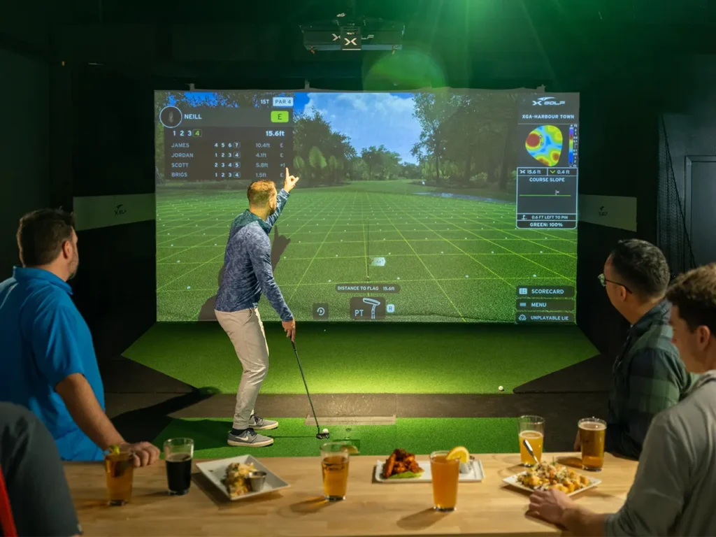 Man pointing to the screen on an indoor golf simulator during a bachelor party at X-Golf, with friends watching from a table filled with drinks and appetizers.