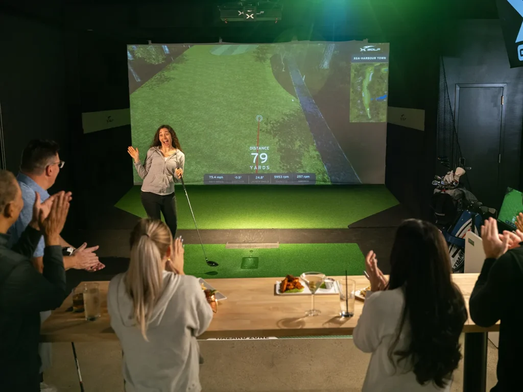 A woman celebrating a successful golf shot during a birthday party at X-Golf, as friends clap and cheer from a nearby table with food and drinks.