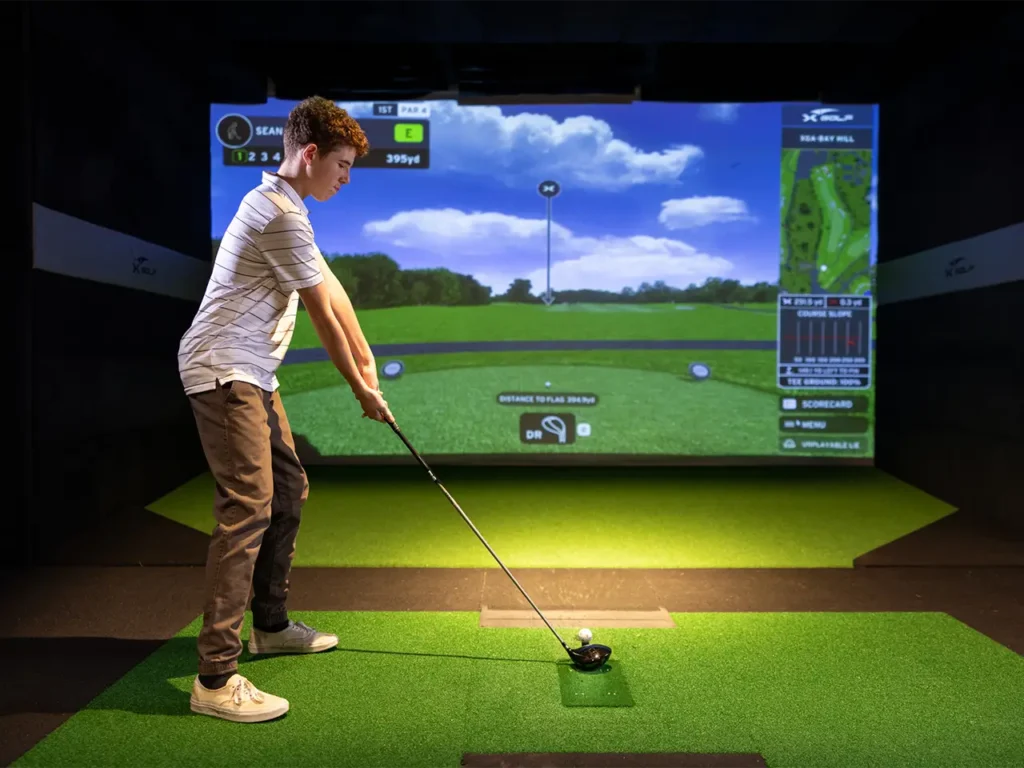 Teenager lining up a golf shot on an indoor simulator at X-Golf during a field trip, with the simulator screen showing a 395-yard hole and course details.