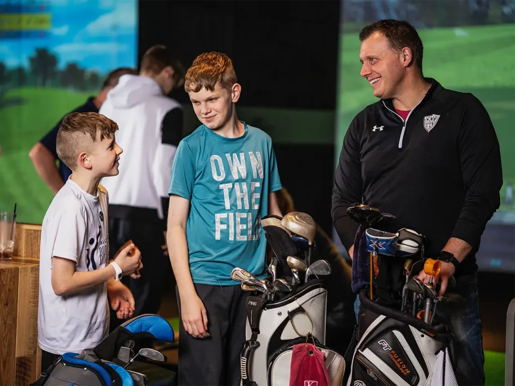 Two boys smiling and chatting with an adult near golf bags during a field trip at X-Golf, with a golf simulator visible in the background.