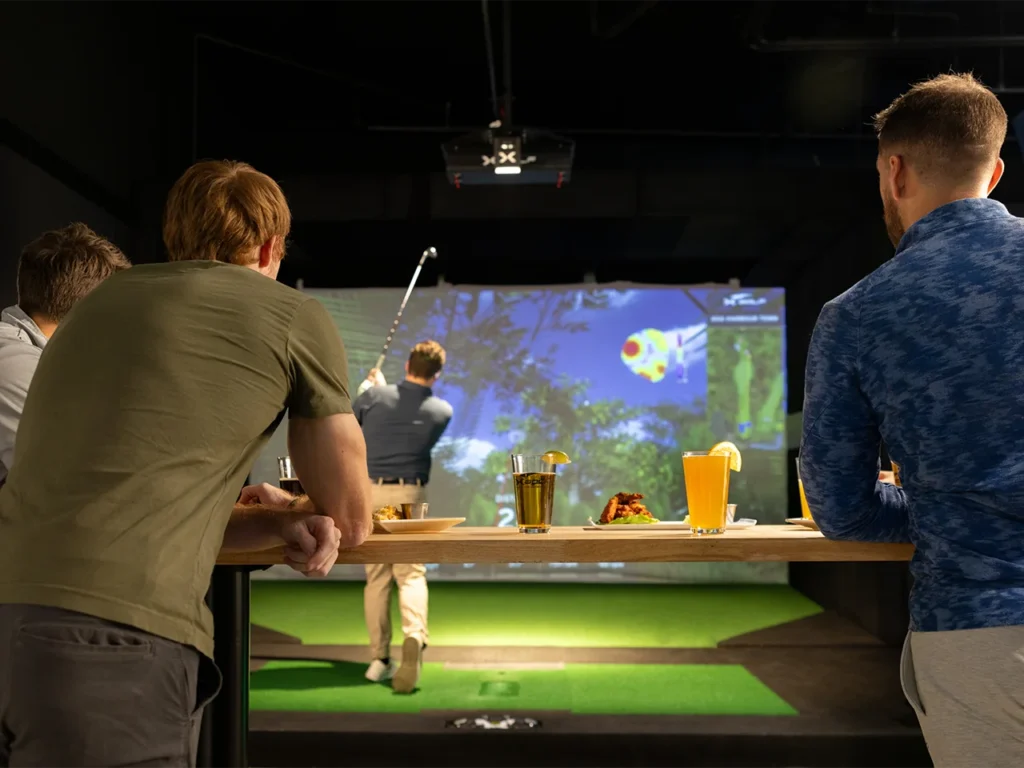 Group of friends enjoying drinks and food while watching a man take a swing at an indoor golf simulator at 'X Golf.'