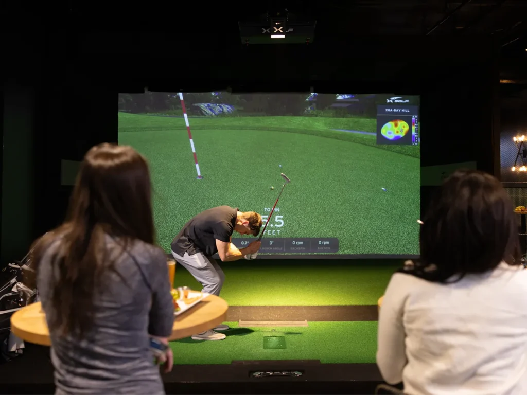 Man lining up a shot on an indoor golf simulator at an 'X Golf' facility, observed by two people holding drinks and appetizers.