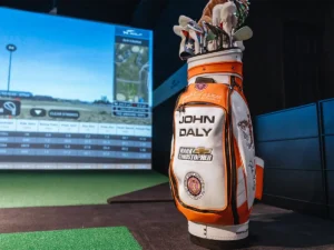 Golf bag with 'John Daly' branding placed in front of a virtual golf simulator screen showcasing a golf course and performance data.