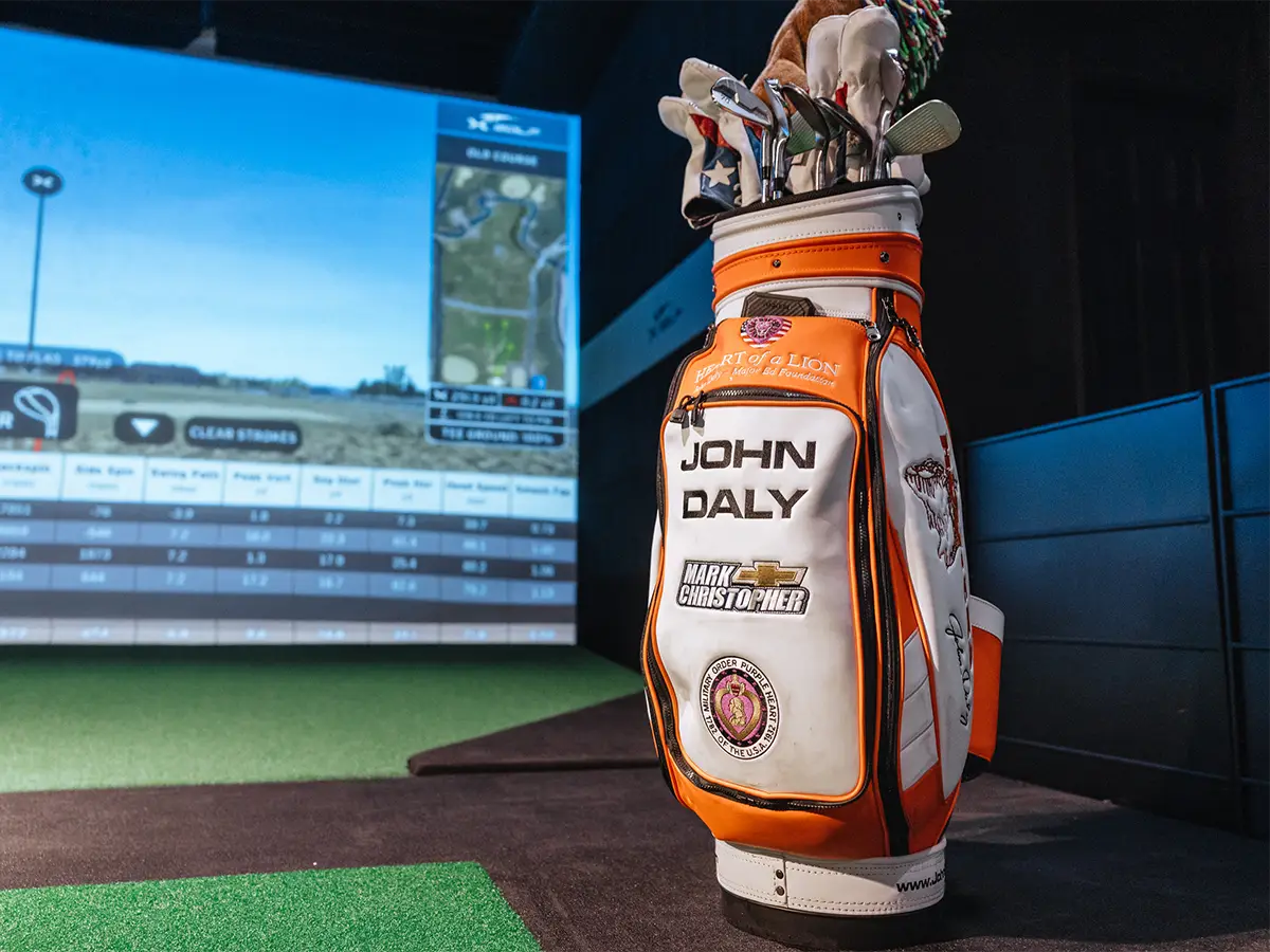 Golf bag with 'John Daly' branding placed in front of a virtual golf simulator screen showcasing a golf course and performance data.