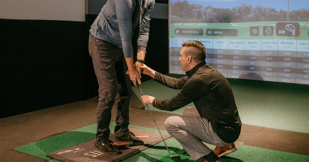 A golf coach crouches down to adjust a student's grip on a club during an indoor lesson at an X-Golf facility, with a simulator displaying shot data.