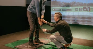 A golf coach crouches down to adjust a student's grip on a club during an indoor lesson at an X-Golf facility, with a simulator displaying shot data.