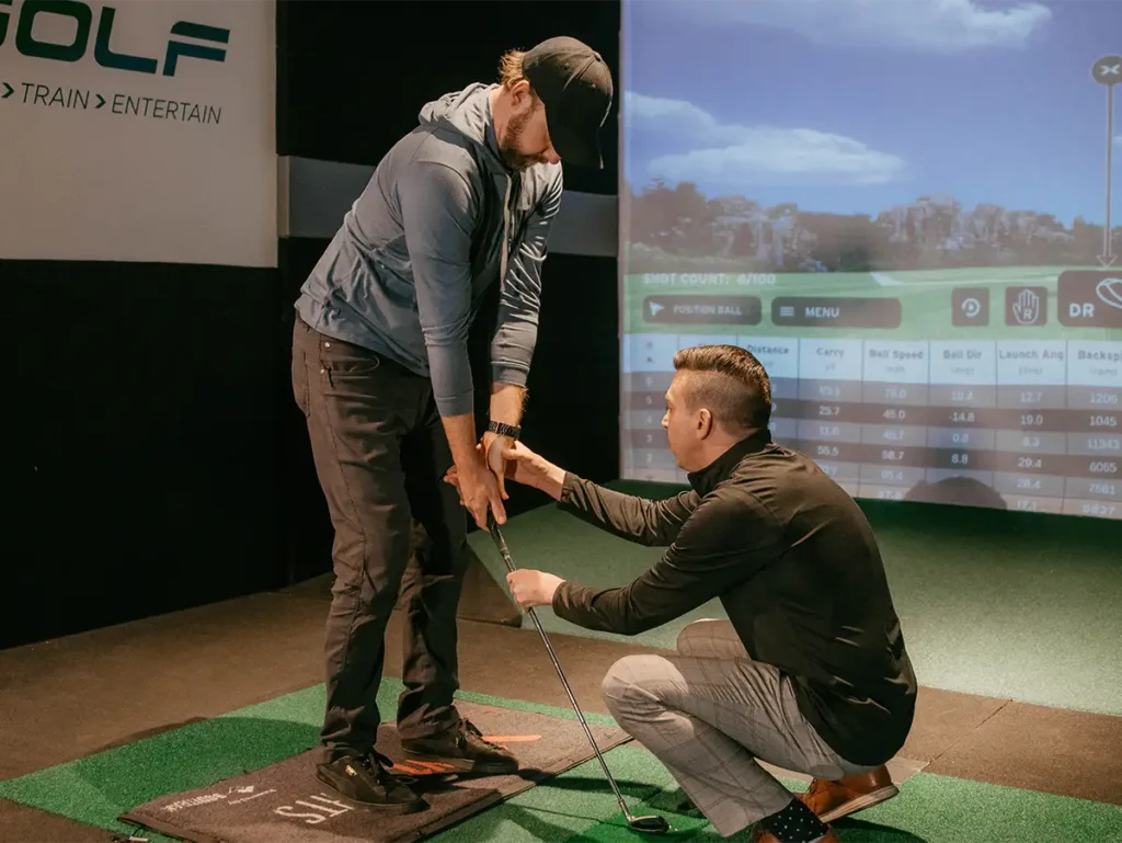 A golf coach crouches down to adjust a student's grip on a club during an indoor lesson at an X-Golf facility, with a simulator displaying shot data.