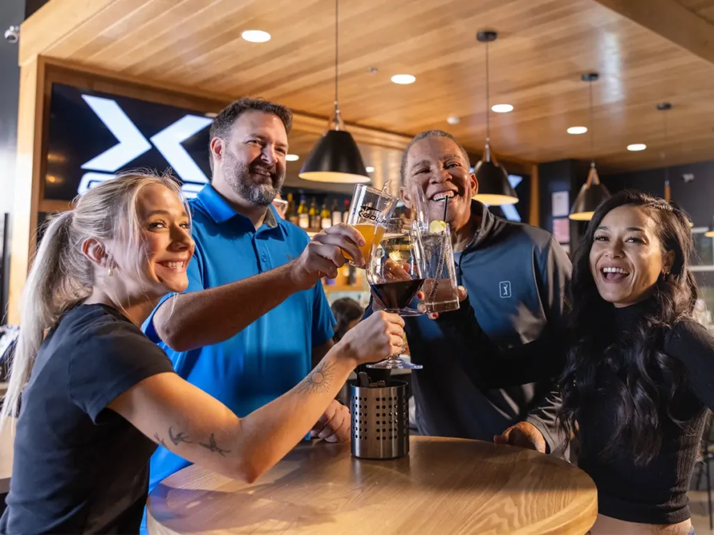 Two couples smiling and raising their glasses in a toast during a double date at X-Golf.