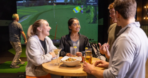 A group of friends enjoys drinks and appetizers around a high-top table at an indoor golf simulator venue. In the background, a large screen displays a virtual golf course while a man takes a swing.