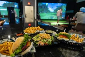 Plates of burgers, fries, chips, and salad on a bar counter with people playing virtual golf in the background.