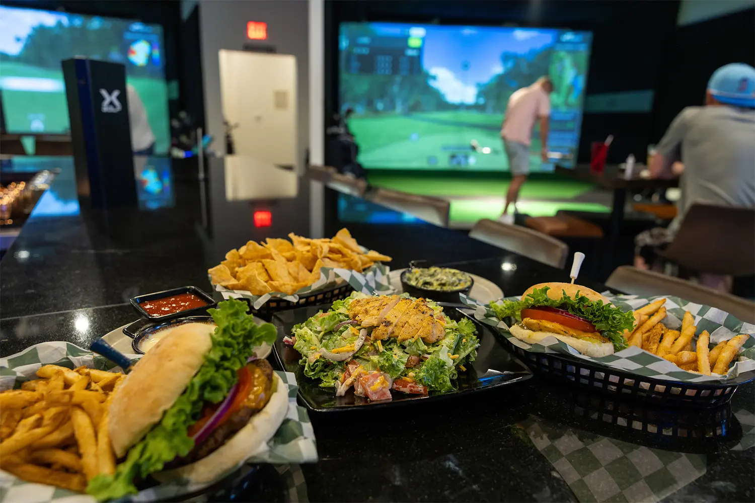 Plates of burgers, fries, chips, and salad on a bar counter with people playing virtual golf in the background.