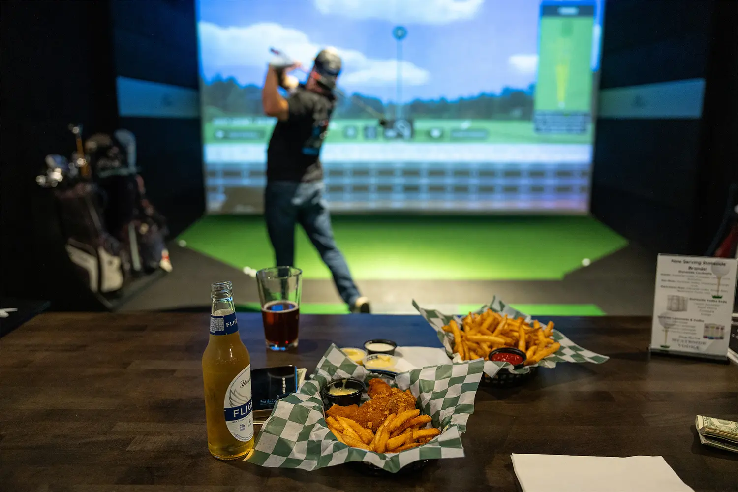 A person swinging a golf club in a simulator while beer and baskets of fries and chicken tenders sit on the table in the foreground.