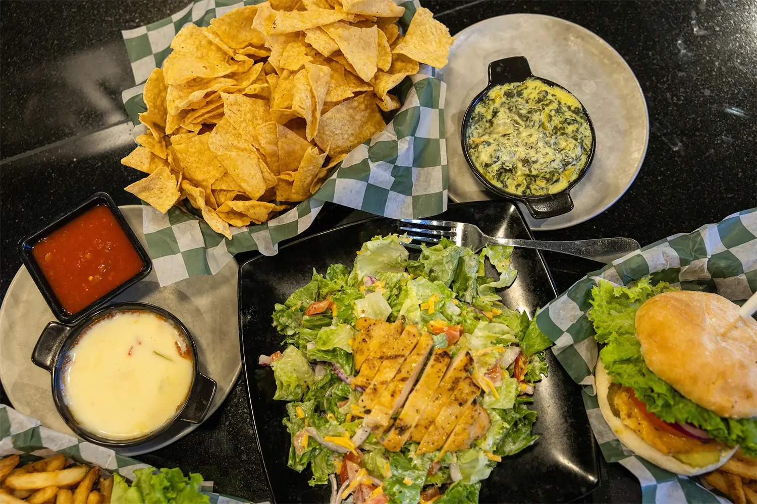 Spread of chips, queso, salsa, salad with grilled chicken, and a burger basket on a black countertop.
