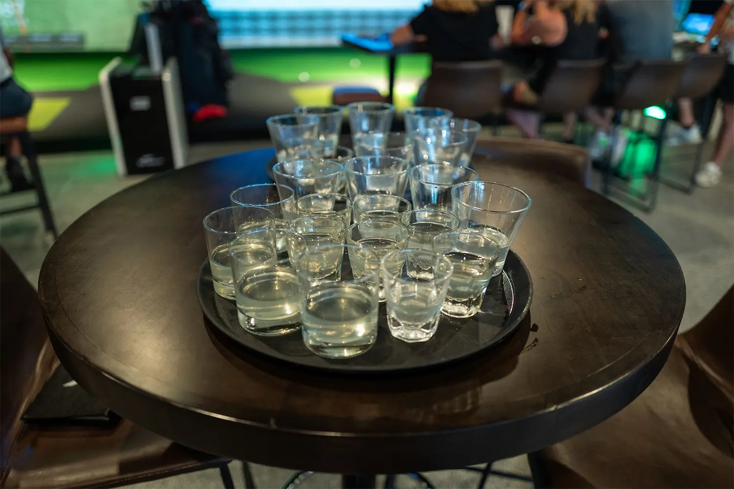 Tray of shot glasses filled with clear liquor on a round table at a golf simulator bar.