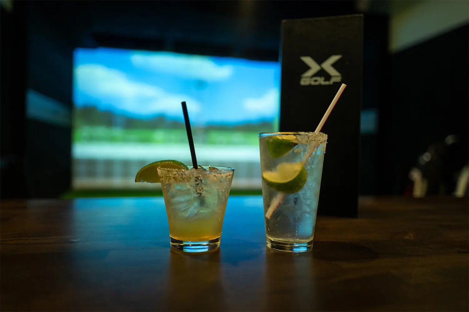 Two cocktails with lime wedges on a table, with a golf simulator screen blurred in the background.
