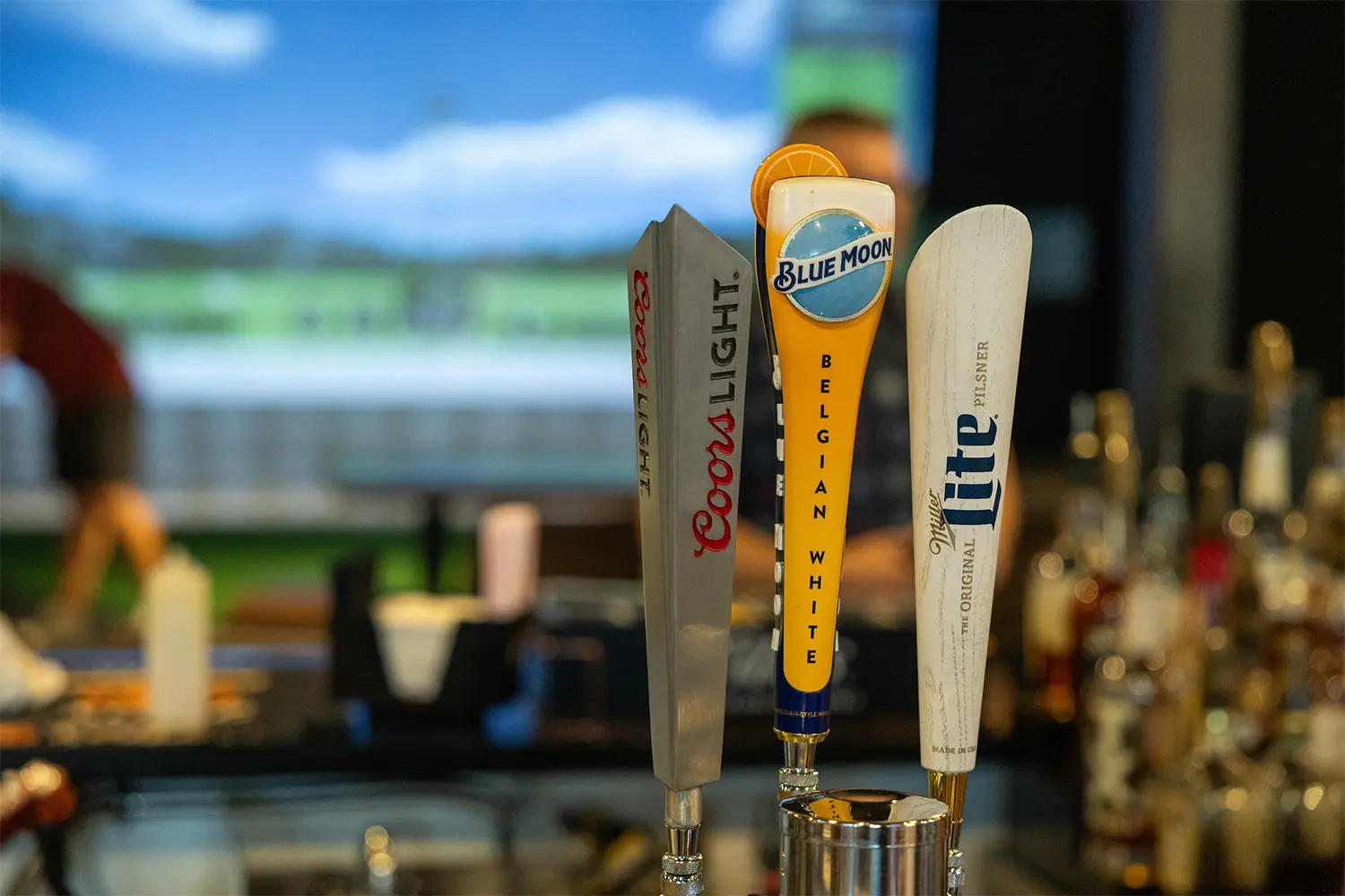 Beer taps for Coors Light, Blue Moon, and Miller Lite at the bar with a golf simulator in the background.