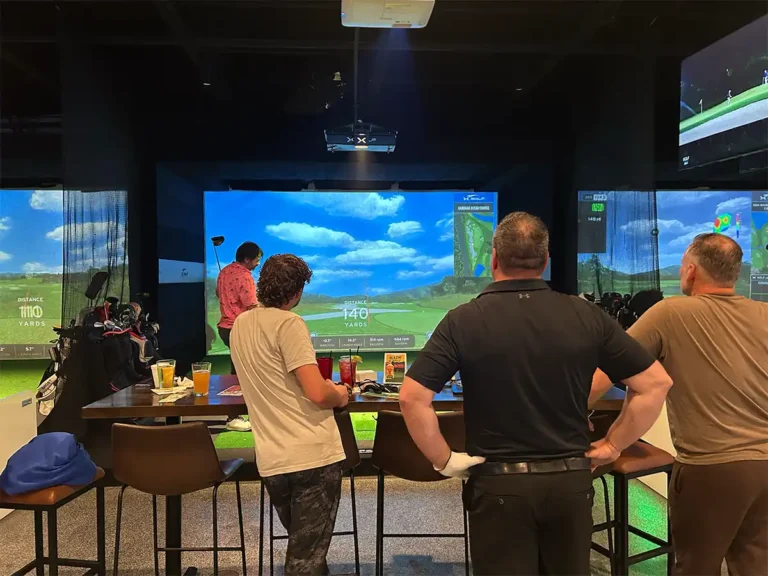 Friends watch league play from bar seating as golfers compete in adjacent simulator bays with drinks on the table.