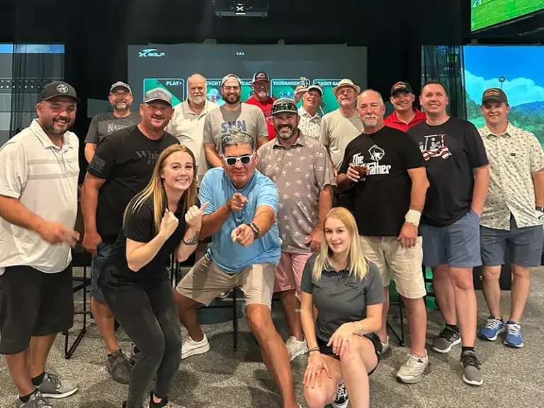 A large group of approximately twenty golfers and two X Golf staff members pose for a group photo in front of an X Golf simulator screen during league night at X Golf Rockwall