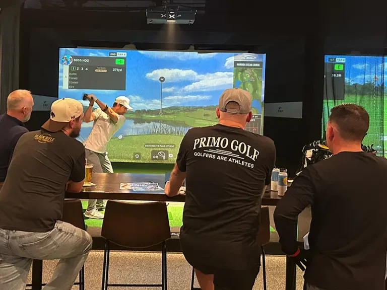 Four men watch a golfer take a tee shot on an indoor simulator screen displaying the Hanbada Ocean Course at X Golf Rockwall, with drinks on the bar counter and a Primo Golf branded shirt visible